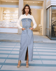 A woman standing in a indoor setting wearing a light blue denim maxi dress with a shirt collar, long sleeves, and button closure. She paired the dress with white strappy sandals.
