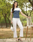 Woman in a white t-shirt, denim vest, and white pants standing outdoors with greenery in the background.